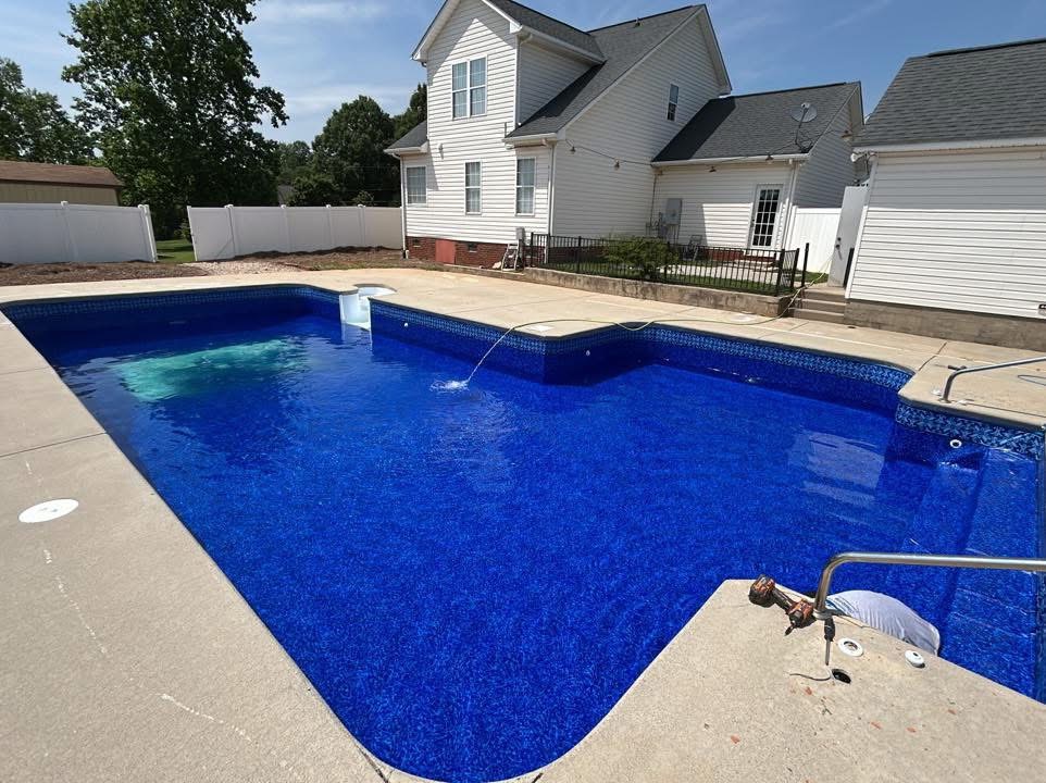 A rectangular backyard swimming pool with clear blue water is surrounded by concrete, next to a white house with a white fence and some green trees in the background.