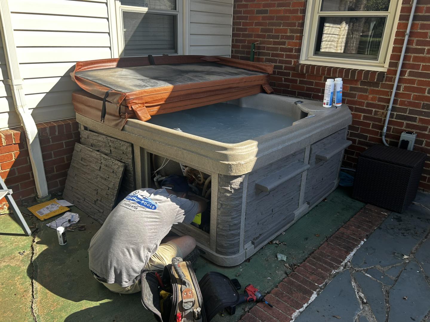 A person kneels beside an outdoor hot tub with the access panel open, performing maintenance. The hot tub cover is partially open, and tools and supplies are nearby.