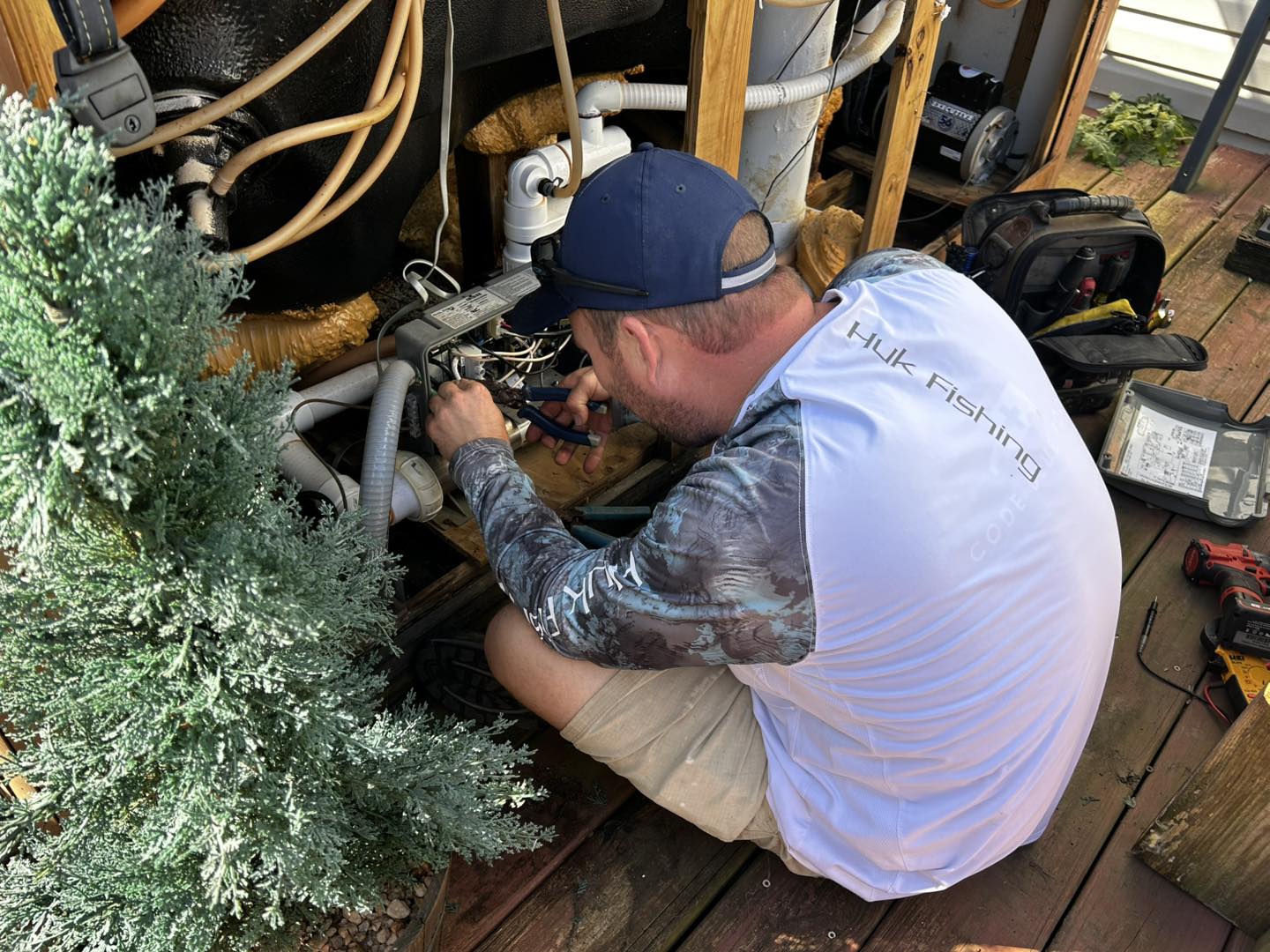 A man in a Huk Fishing shirt repairs or installs electrical equipment among pipes on a wooden deck, with tools and a small tree nearby.