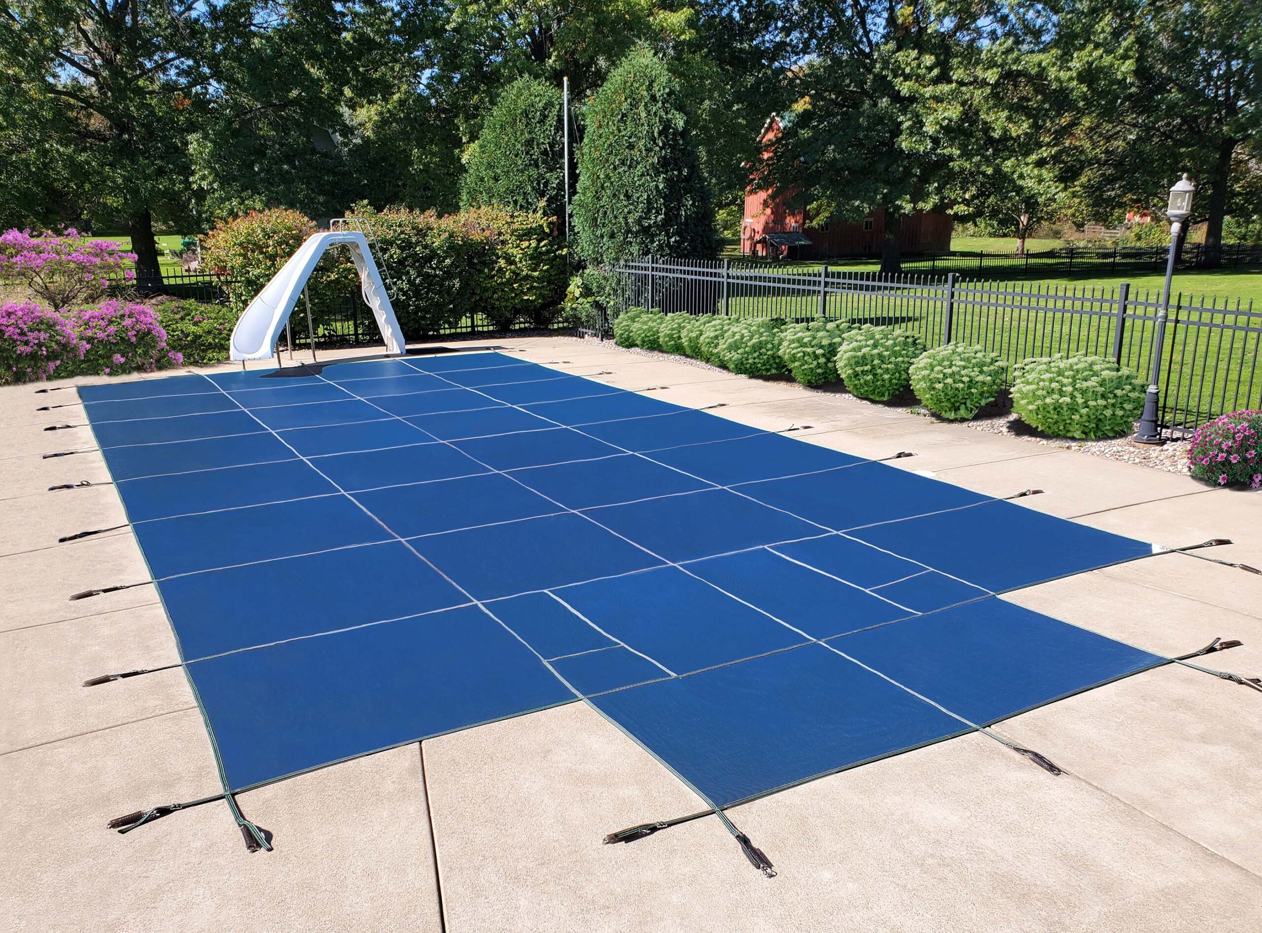 A rectangular in-ground pool is covered with a blue safety cover secured by straps; a white slide and black metal fence are visible in a landscaped backyard.