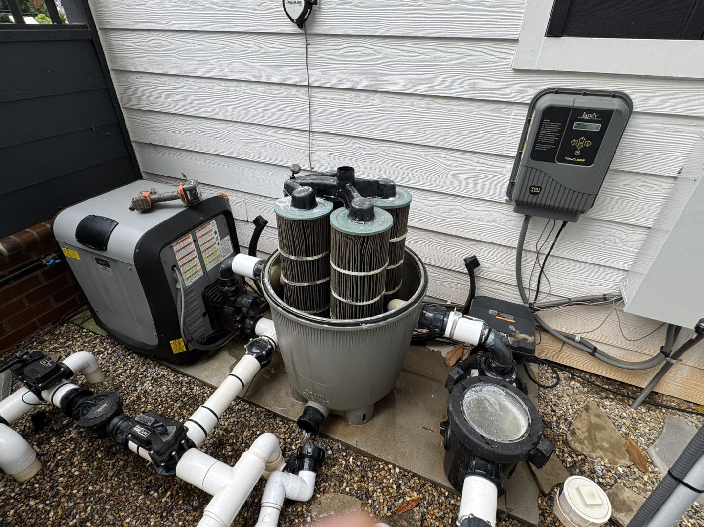 A pool filter with exposed cartridges is shown outside next to a heater, control box, and various pipes on a gravel surface beside a white building.