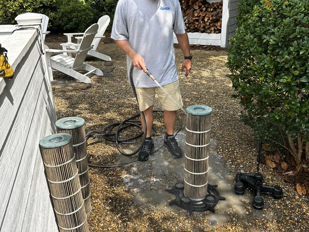 A person uses a hose to clean cylindrical pool filters outdoors. Two clean filters stand upright, while another lies on the ground next to filtering equipment.