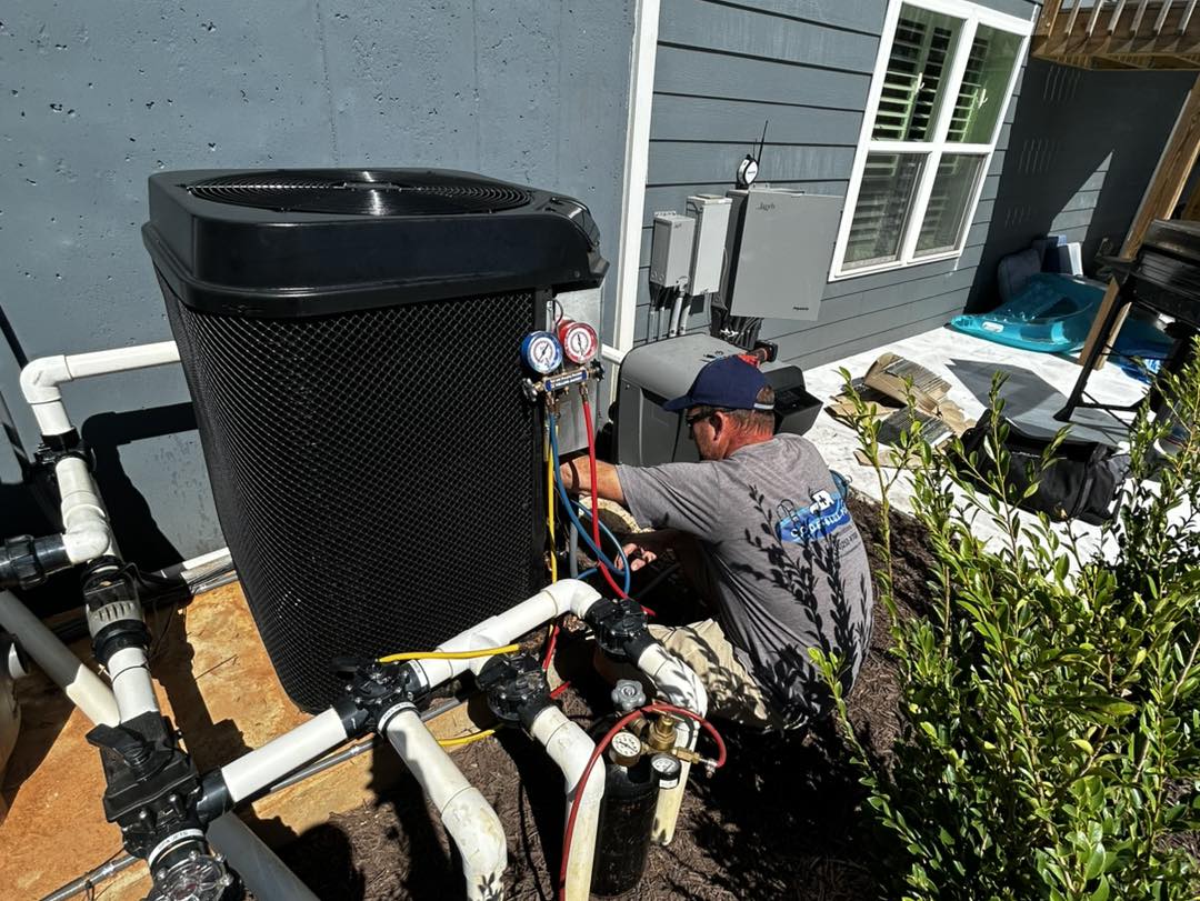 A technician in a gray shirt services an outdoor HVAC unit beside a house, using gauges to check system pressure and connected pipes.