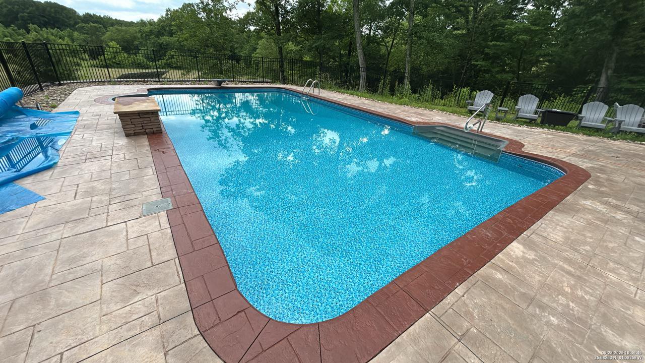 Rectangular outdoor swimming pool with a red-tiled border, surrounded by stamped concrete patio and Adirondack chairs, set near trees and a black metal fence.