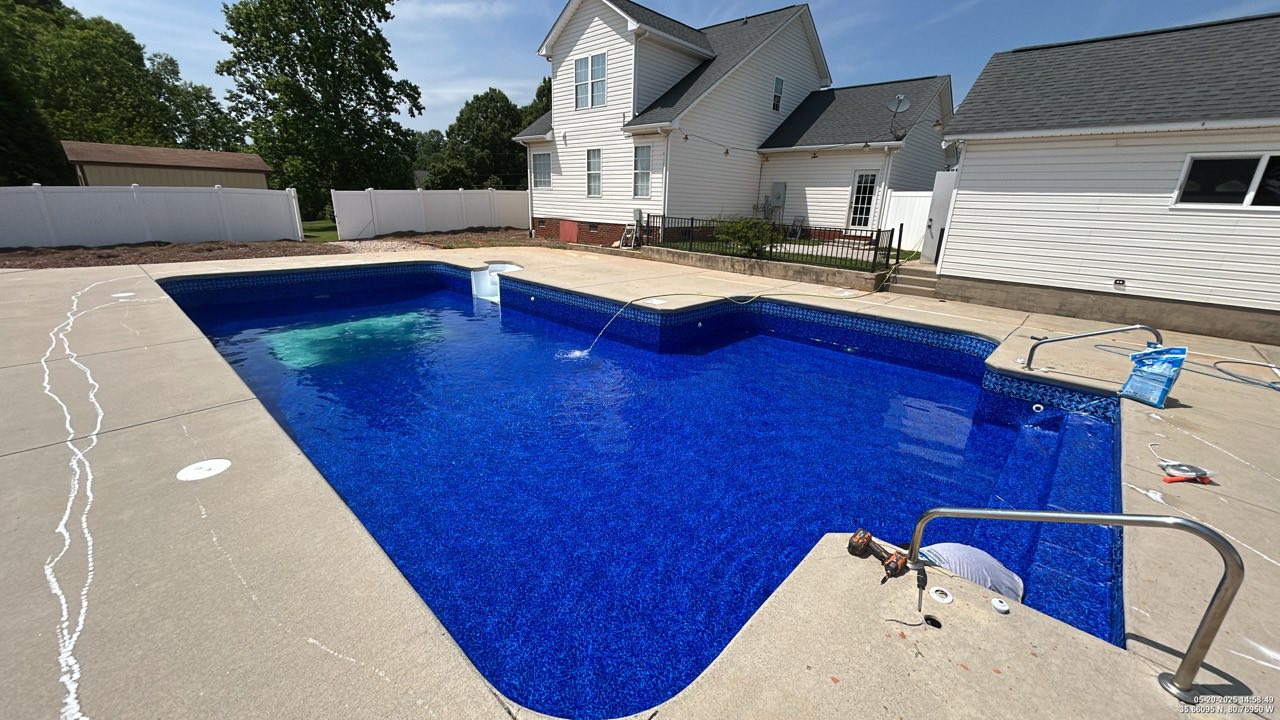 A rectangular in-ground swimming pool with blue water is surrounded by a concrete patio in the backyard of a white house on a sunny day.