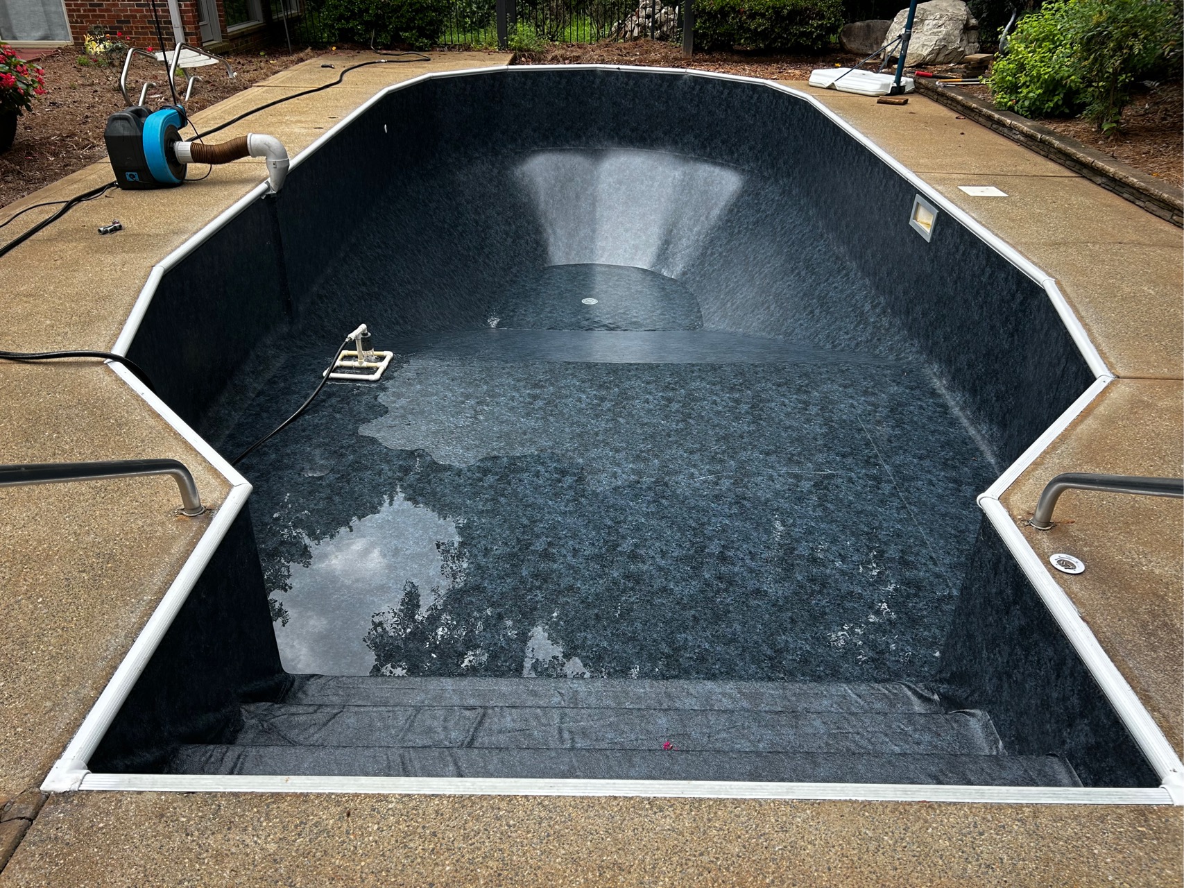 Empty in-ground swimming pool with a dark liner, some water at the bottom, a ladder on the left, and pool maintenance equipment nearby on the concrete pool deck.