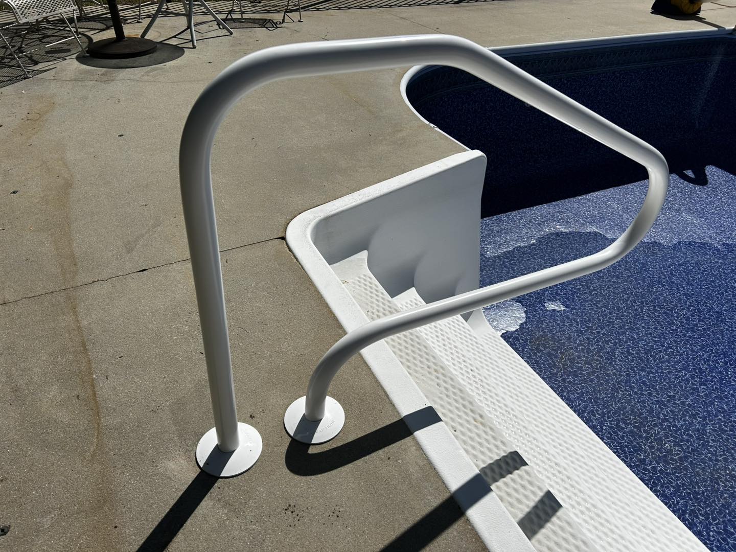 A white metal handrail is installed by the edge of an outdoor swimming pool with a concrete deck and pool steps leading into the water.