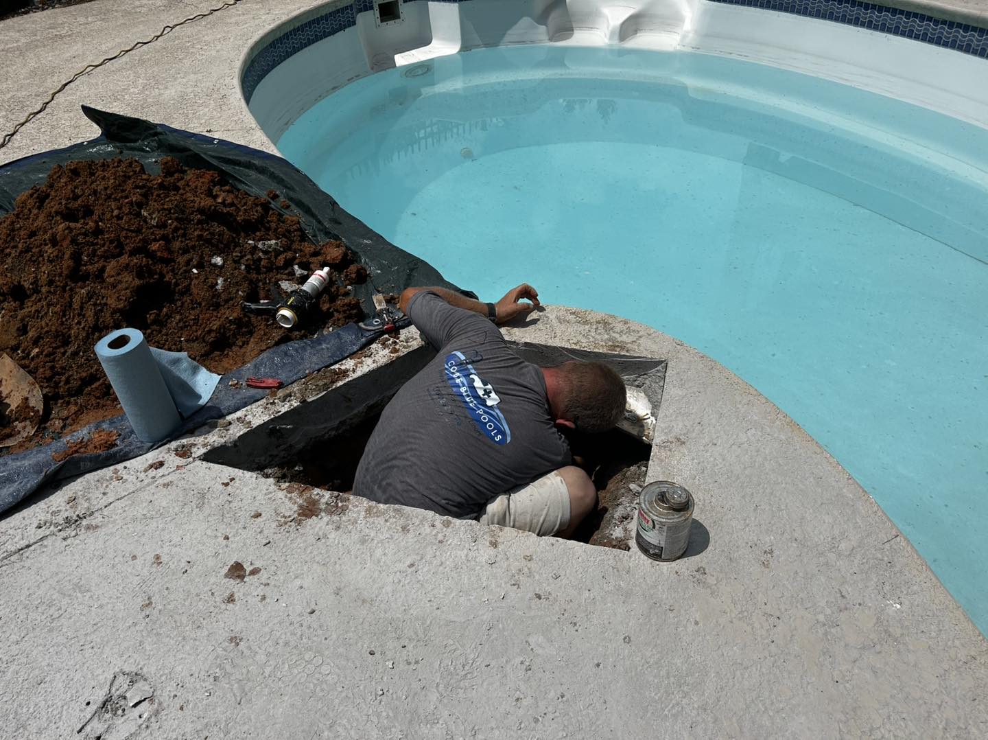 A worker kneels in a cut-out section of concrete beside a swimming pool, performing maintenance or repairs with tools and materials nearby.