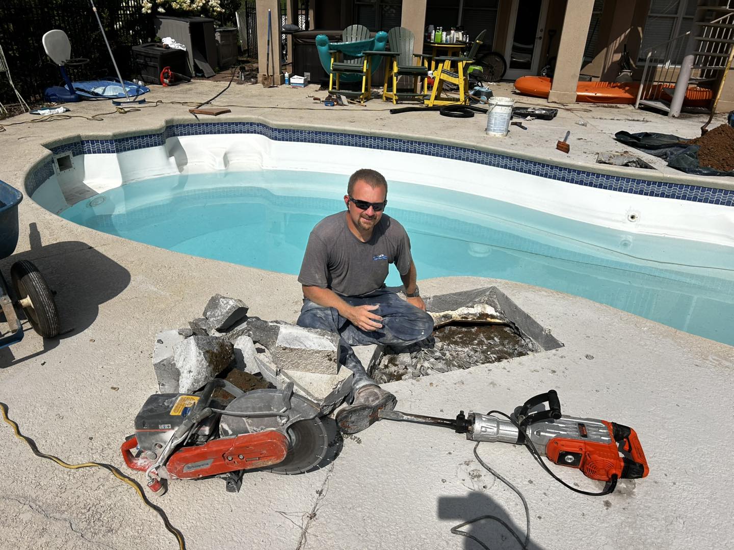 A man kneels on a pool deck beside broken concrete and power tools, with a hole cut into the deck near the swimming pool.